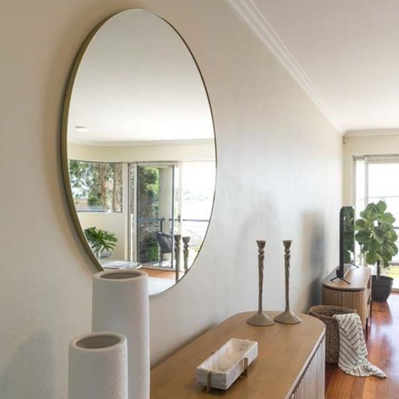 A room with a large oval mirror, a wooden console table with decorative items, and a view of a balcony with outdoor plants.