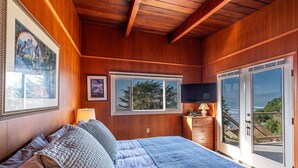 Un dormitorio con una cama grande, paredes de madera, un televisor, un aparador y vistas al océano a través de puertas de vidrio.
