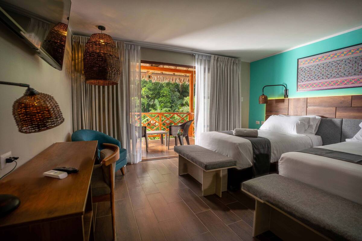 A hotel room with two beds, a wooden desk, a chair, and a balcony with a view of greenery.