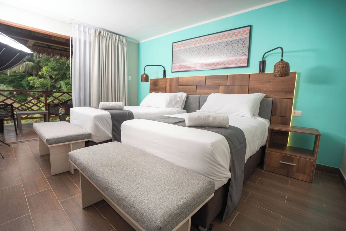 A hotel room with two beds, a bench, a nightstand, and a view of greenery outside.