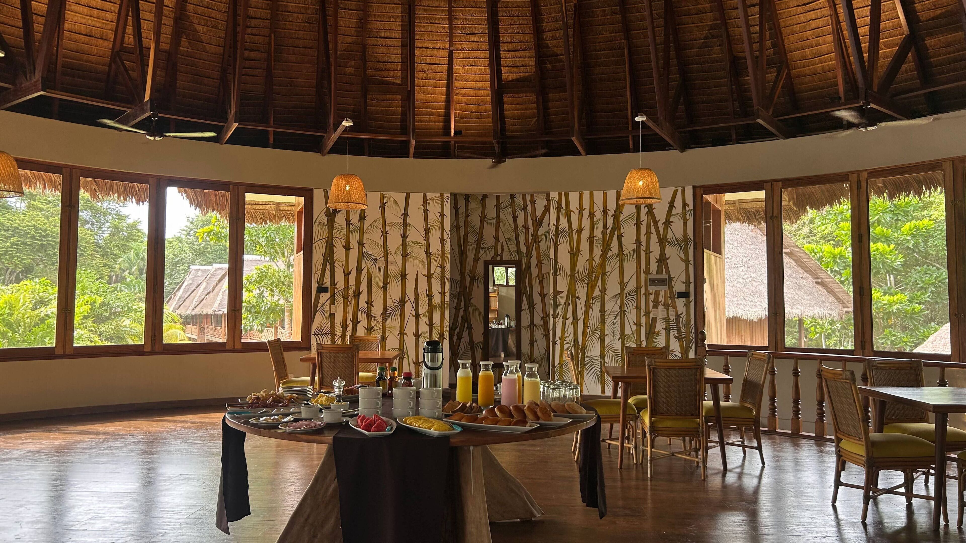 A dining area with a large round table set for breakfast, surrounded by chairs, with a view of greenery outside.