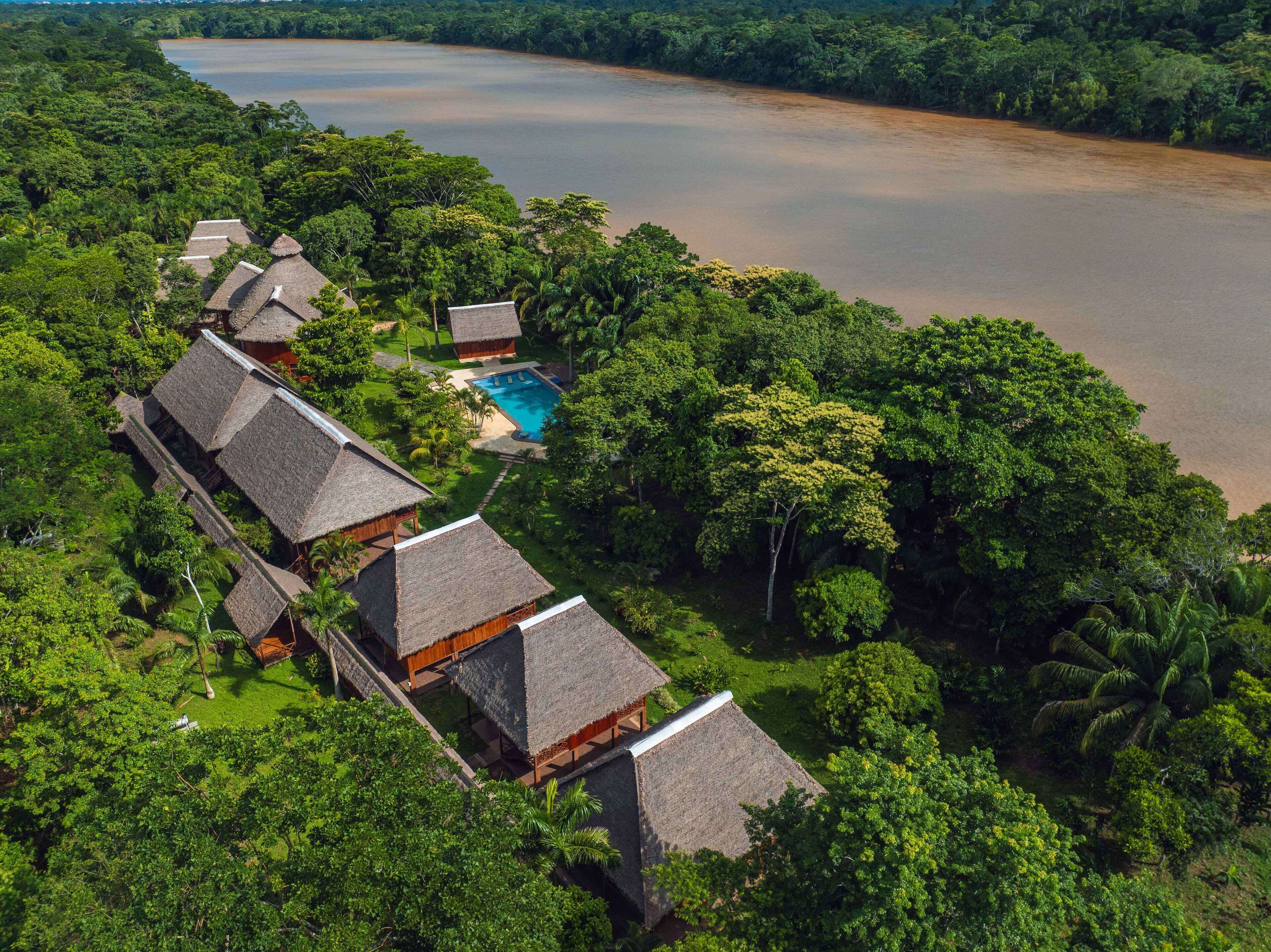 An aerial view of a resort with thatched-roof bungalows, a swimming pool, and a large body of water.