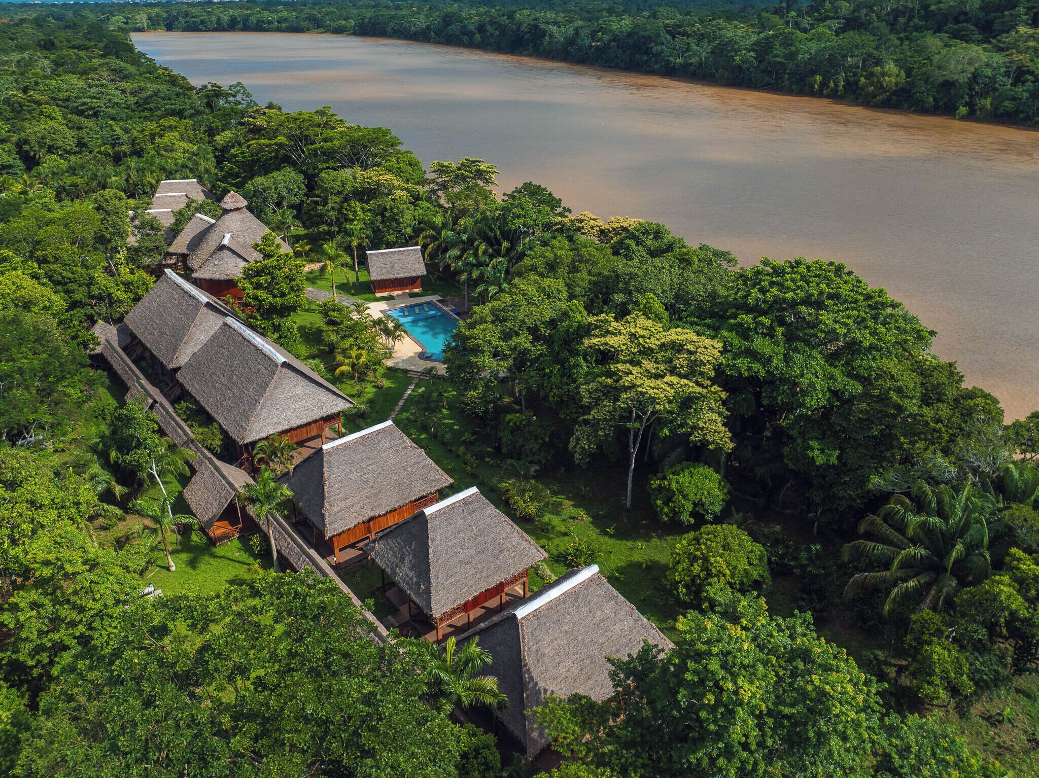 An aerial view of a resort with thatched-roof villas, a swimming pool, and a large body of water.