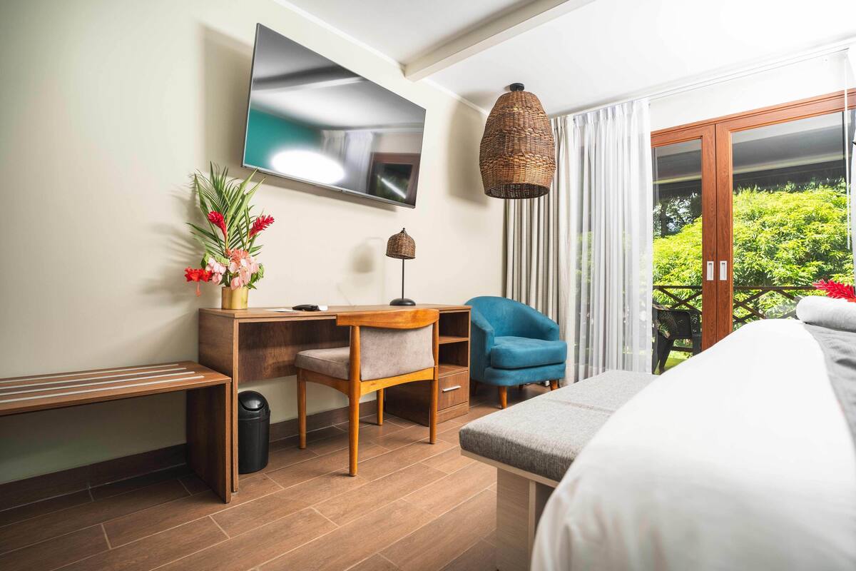 A modern hotel room with a flat-screen TV, a wicker pendant light, a desk with a chair, a blue armchair, and a bed.