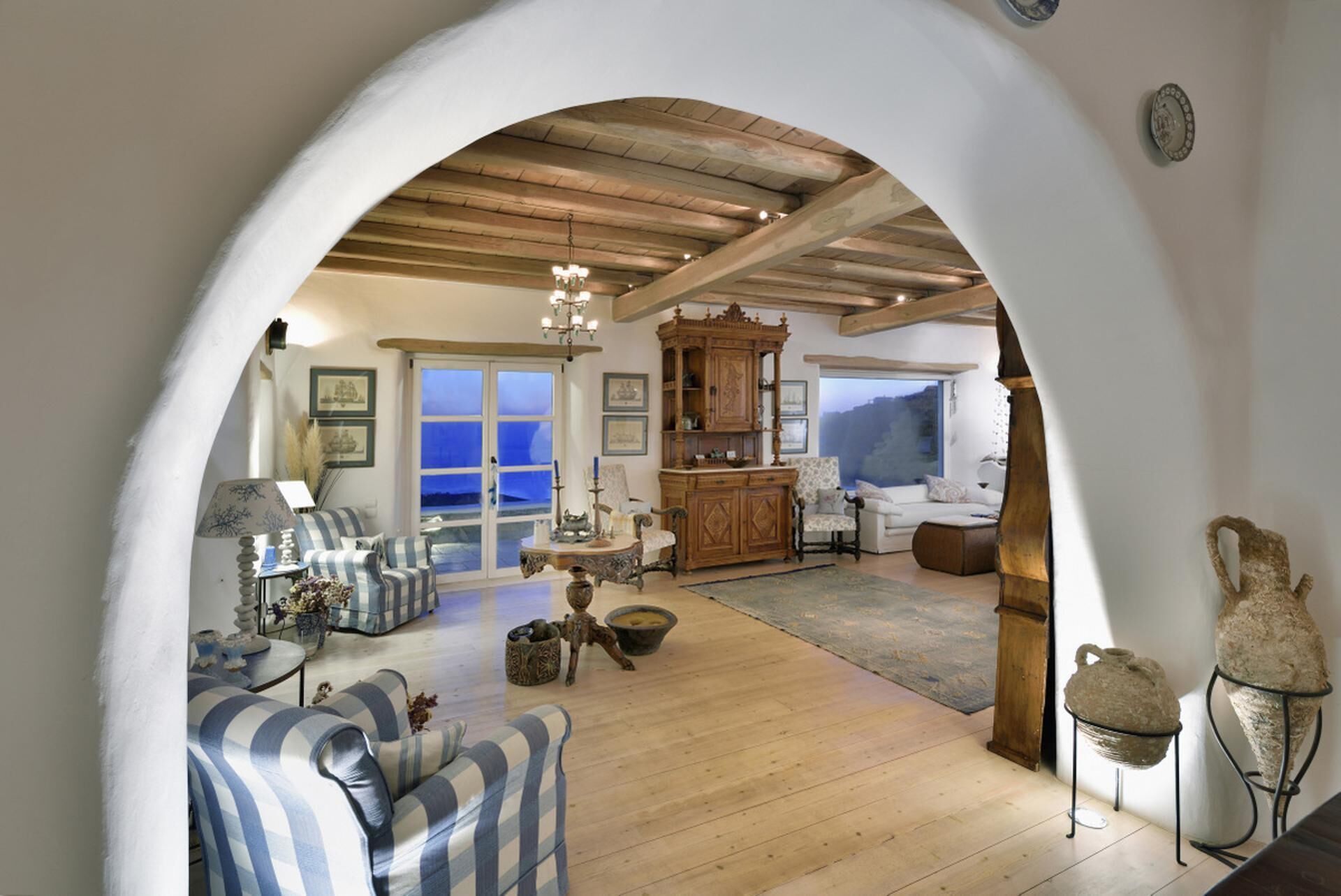 Un salón espacioso con techo alto, vigas de madera y un amplio arco que da a un balcón. Cuenta con varias zonas de estar, un aparador de madera y vistas al exterior.