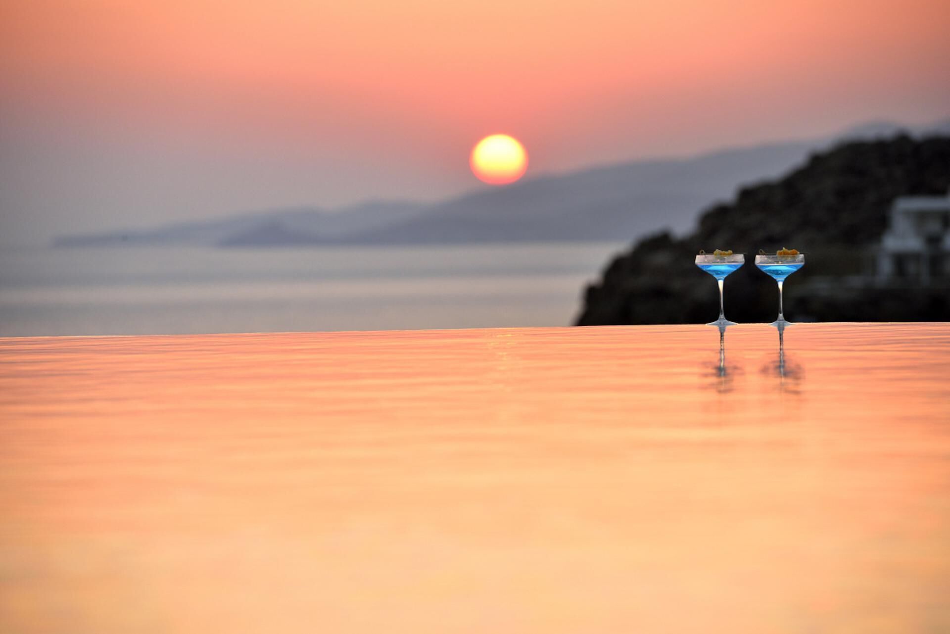 Dos vasos de cóctel sobre una superficie de madera, con un atardecer y un mar tranquilo al fondo.