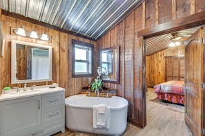 A wooden interior with a bathtub, a vanity with a mirror, and a bed with a floral bedspread. - Converted Barn Couple's Oasis Near Tallulah Gorge (Clarkesville)
