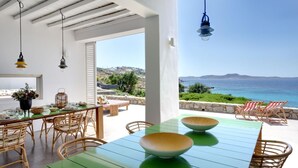 A dining area with a view of the sea, wicker furniture, and hanging lights. - Stunning 5-bed - Villa Big Blue (Mykonos)