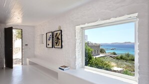 A room with a large window offering a view of the sea and a stone building. There are two framed artworks on the wall. - Stunning 5-bed - Villa Big Blue (Mykonos)