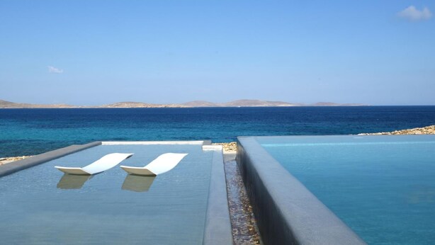 An infinity pool with two white lounge chairs, a clear blue sky, and a view of the sea and distant land. - Stunning 5-bed - Villa Big Blue (Mykonos)