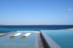 Uma piscina infinita com duas cadeiras de praia brancas, céu azul límpido e vista para o mar e terra distante.