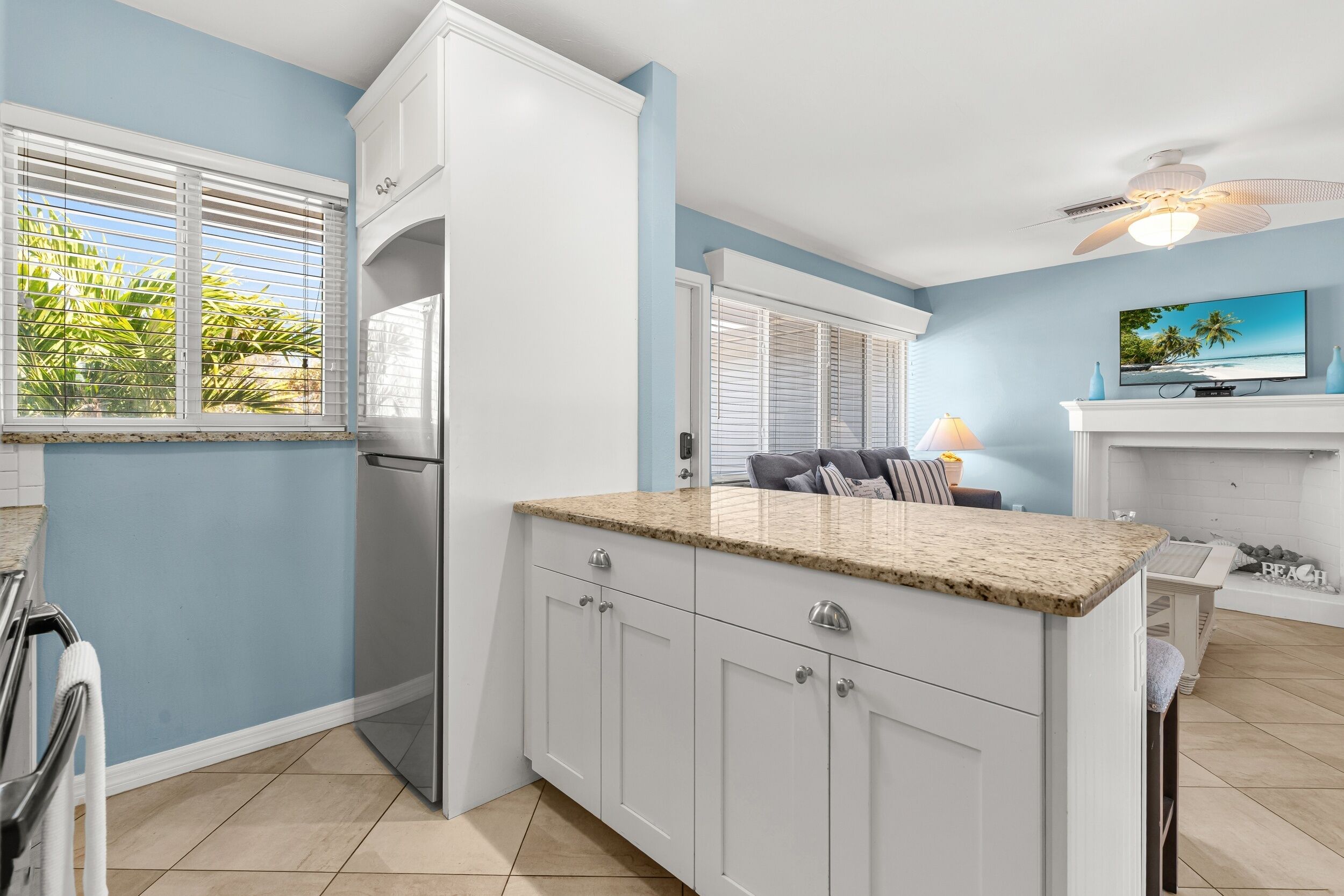 A kitchen with white cabinets, a granite countertop, and a built-in island. There is a window with blinds, a ceiling fan, and a television on the wall.