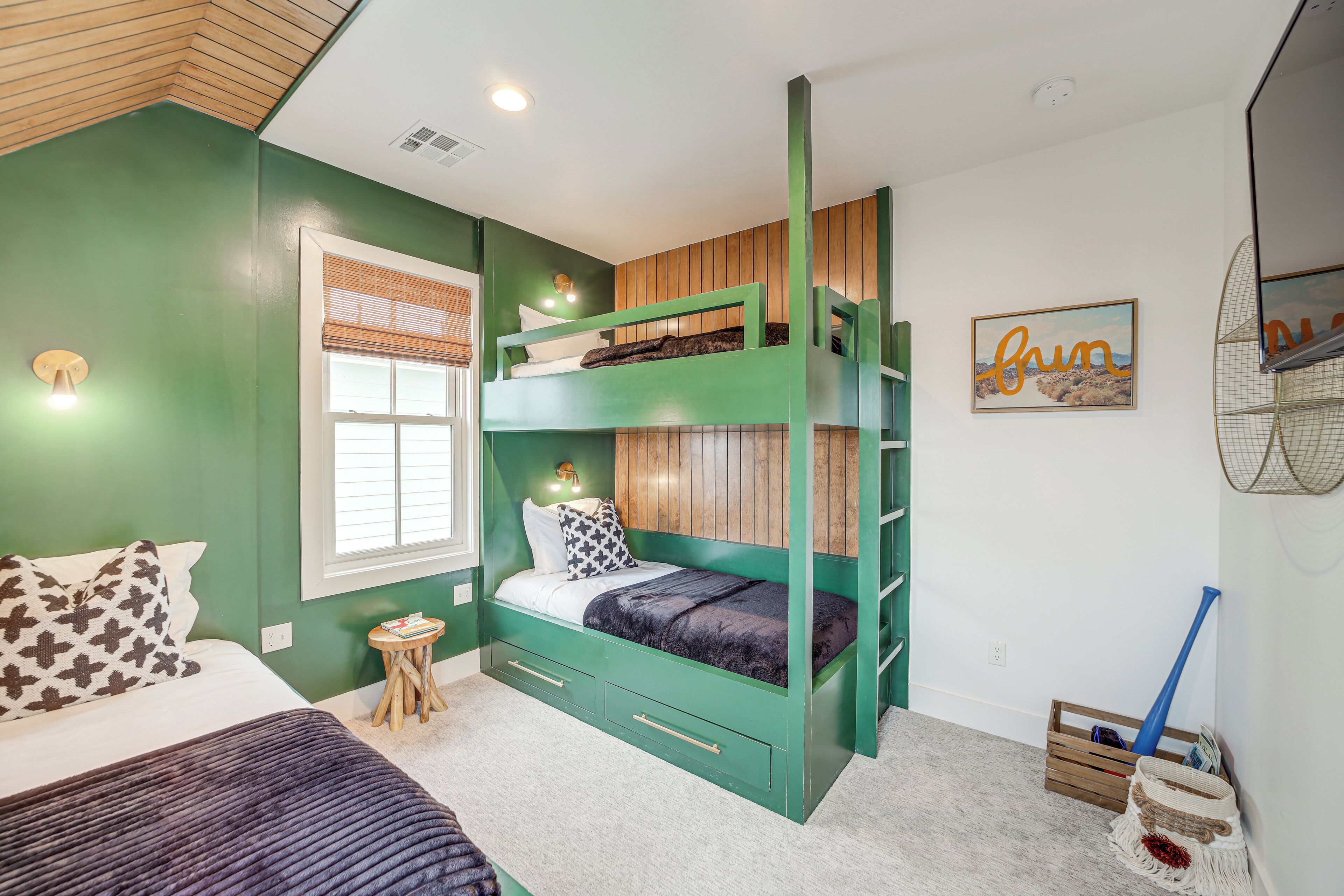 Quarto com beliche, uma mesinha de apoio, janela com persianas e um quadro na parede.