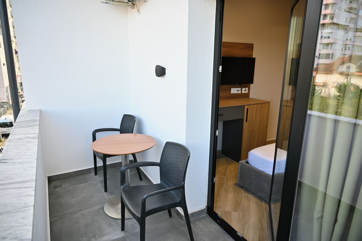 A balcony with a table and chairs, a wooden cabinet, and a bed inside a room.