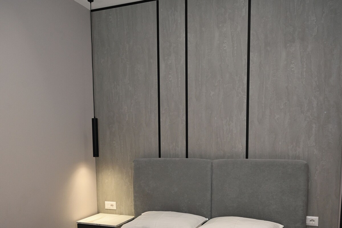 Ein modernes Schlafzimmer mit einem grau gepolsterten Bett, einem Nachttisch mit Lampe und einer Wandleuchte.