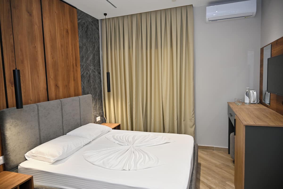 A hotel room with a bed, wooden wardrobe, curtains, a desk with a mirror, and an air conditioning unit.