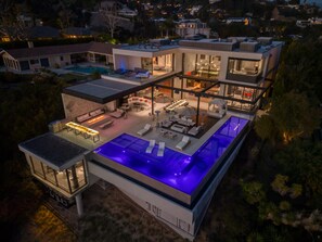 Una casa moderna de dos pisos con piscina en la azotea, mobiliario exterior y un amplio frente de cristal.