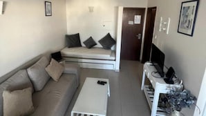 A compact living space with a sofa, a small coffee table, a bed with pillows, a television, and a white cabinet with a vase. - 1-bed - Sleeps 4 - Free Parking (Portimão)