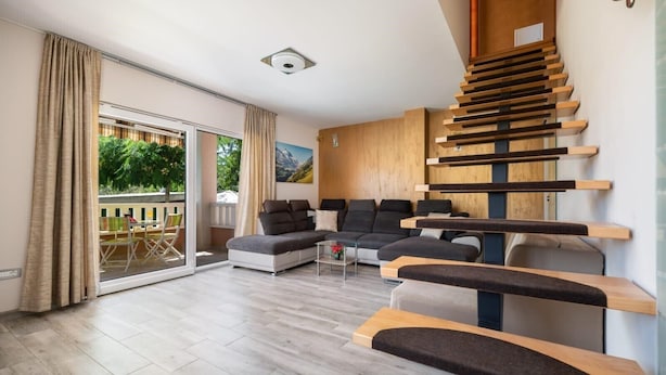 A modern living room with a sectional sofa, a glass door leading to an outdoor area, and a unique wooden staircase.