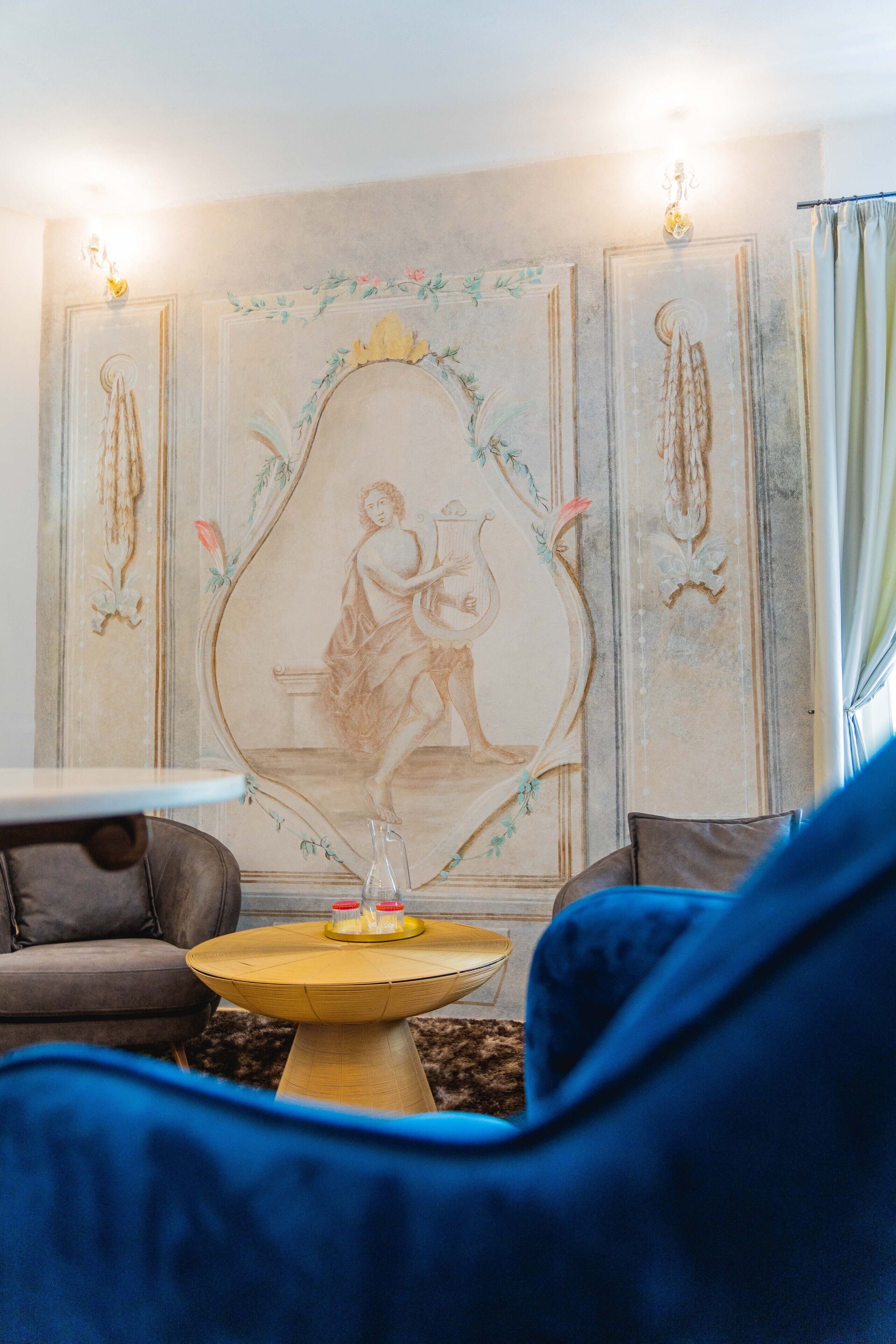 A room with a sofa, a small round table with bottles, and a wall with a classical painting.