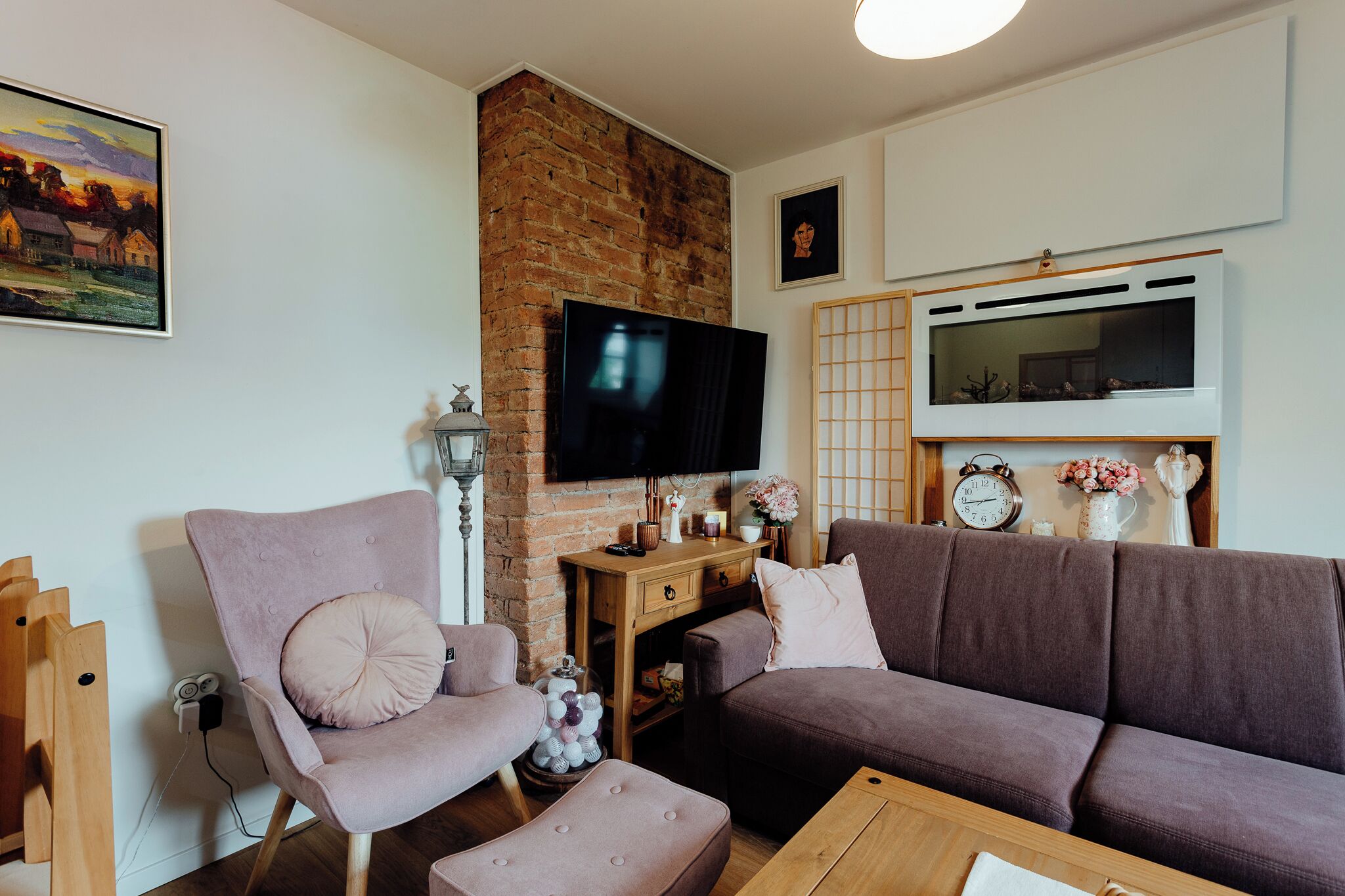 A living room with a brick wall, a flat-screen TV, a sofa, a chair, a wooden coffee table, and a small side table with decorative items.