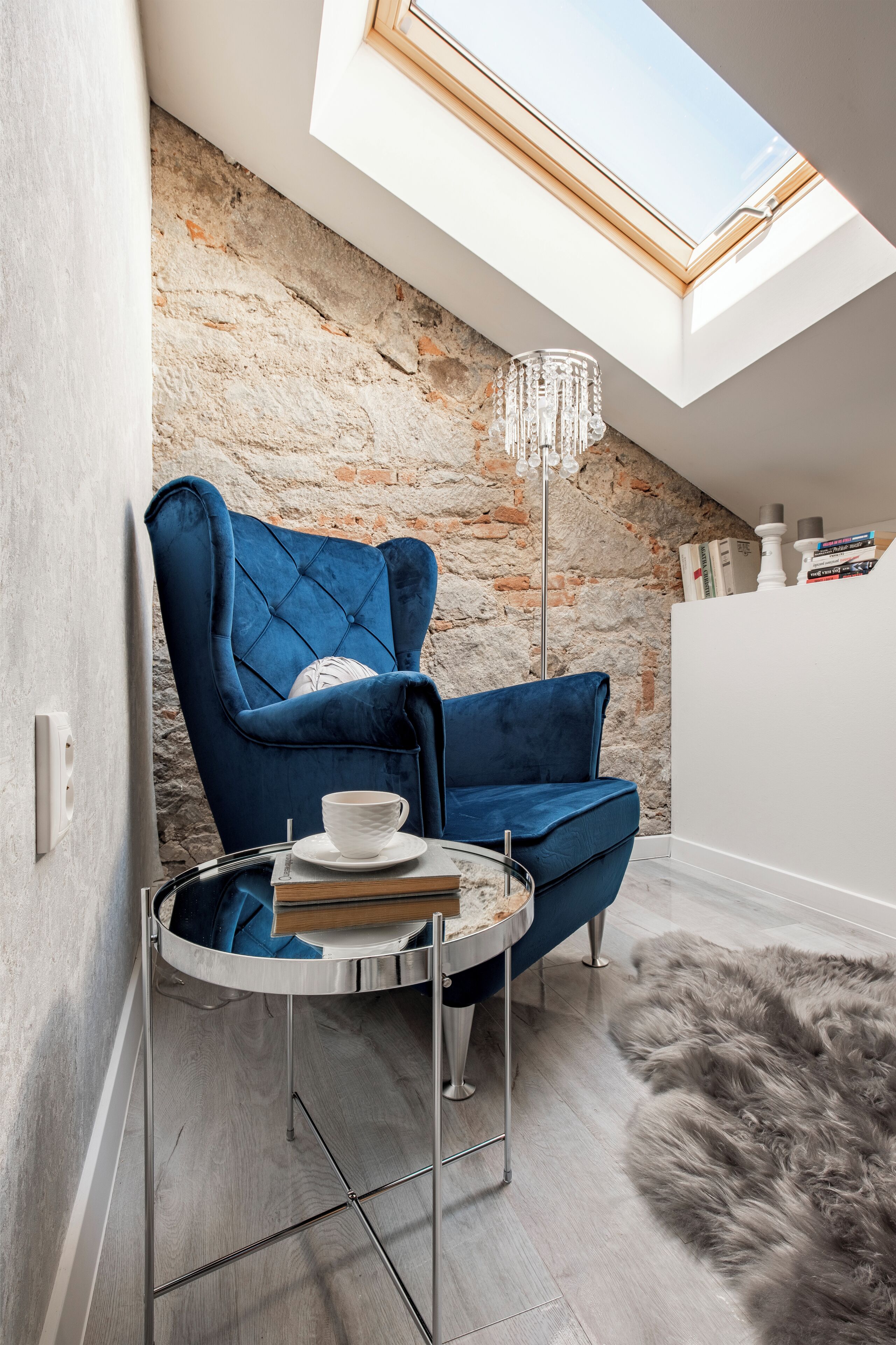 Una habitación acogedora con un sillón de terciopelo azul, una mesita redonda con una taza y libros, un tapete mullido y una pared de piedra con tragaluces.