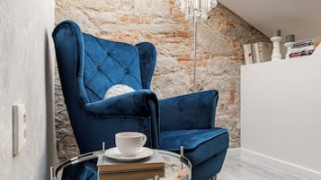 Una habitación acogedora con un sillón de terciopelo azul, una mesita redonda con una taza y libros, un tapete mullido y una pared de piedra con tragaluces.