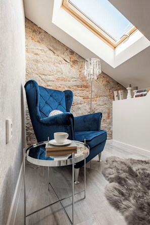 A cozy room with a blue velvet armchair, a small round table with a cup and books, a fluffy rug, and a stone wall with a skylight. - Opera apartments  (Banska Stiavnica)