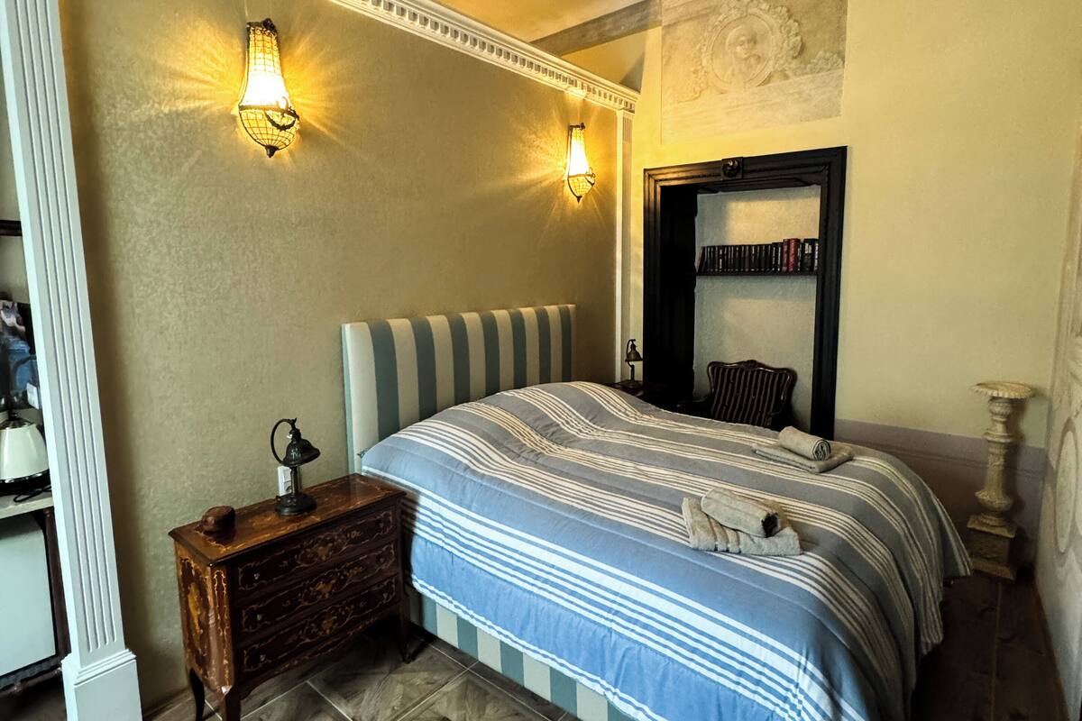 A bedroom with a striped bedspread, two wall sconces, a nightstand with a lamp, a dresser, and a mirror above the bed.