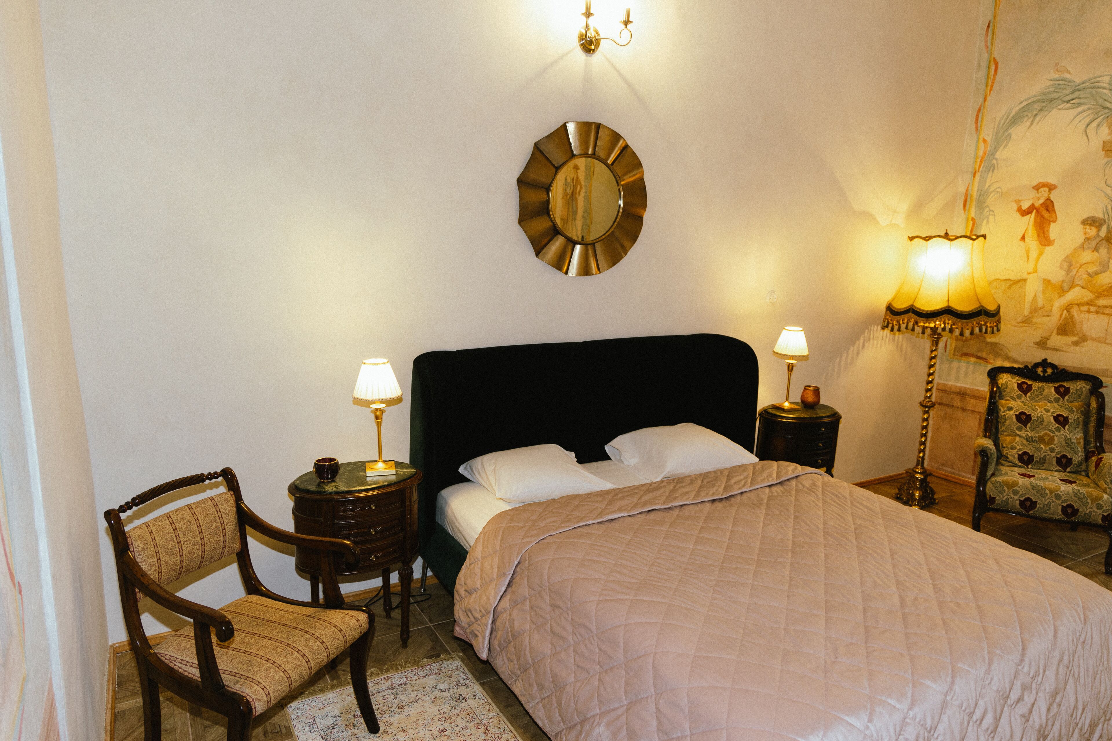 A neatly made bed with a dark headboard, two bedside lamps, a chair, and a wall-mounted mirror.