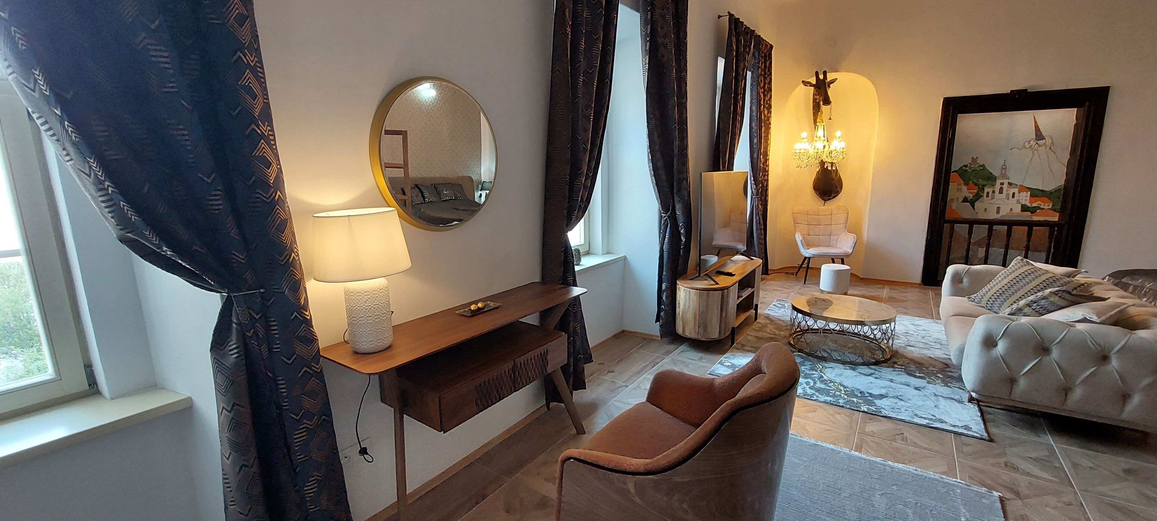 Een moderne woonkamer met een bank, fauteuil en een kleine ronde tafel. Er is een houten bureau met een lamp en een spiegel aan de muur. Een kroonluchter hangt boven de zithoek.