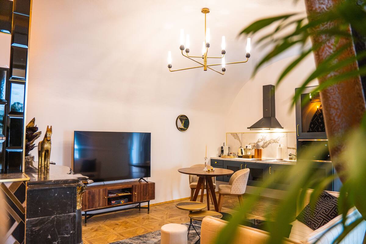 A modern living room with a flat-screen TV, a wooden entertainment unit, a round dining table with chairs, and a kitchen area with a stove and cabinets.