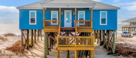 A blue beach house with a wooden deck and a door, elevated on stilts, with a view of the beach and ocean.