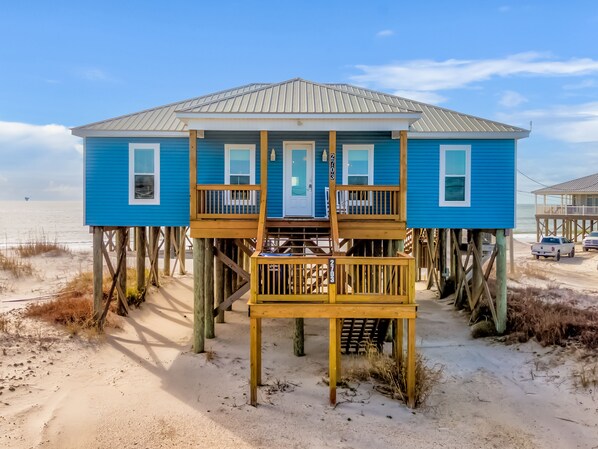 A blue beach house with a wooden deck and a door, elevated on stilts, with a view of the beach and ocean.