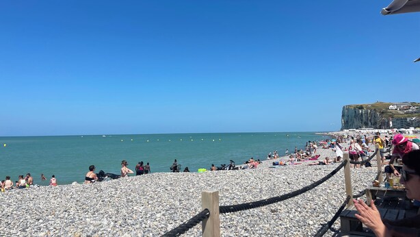Ein Strand mit badenden Personen, einem felsigen Ufer und einem Gebäude im Hintergrund, das auf einer Klippe steht.