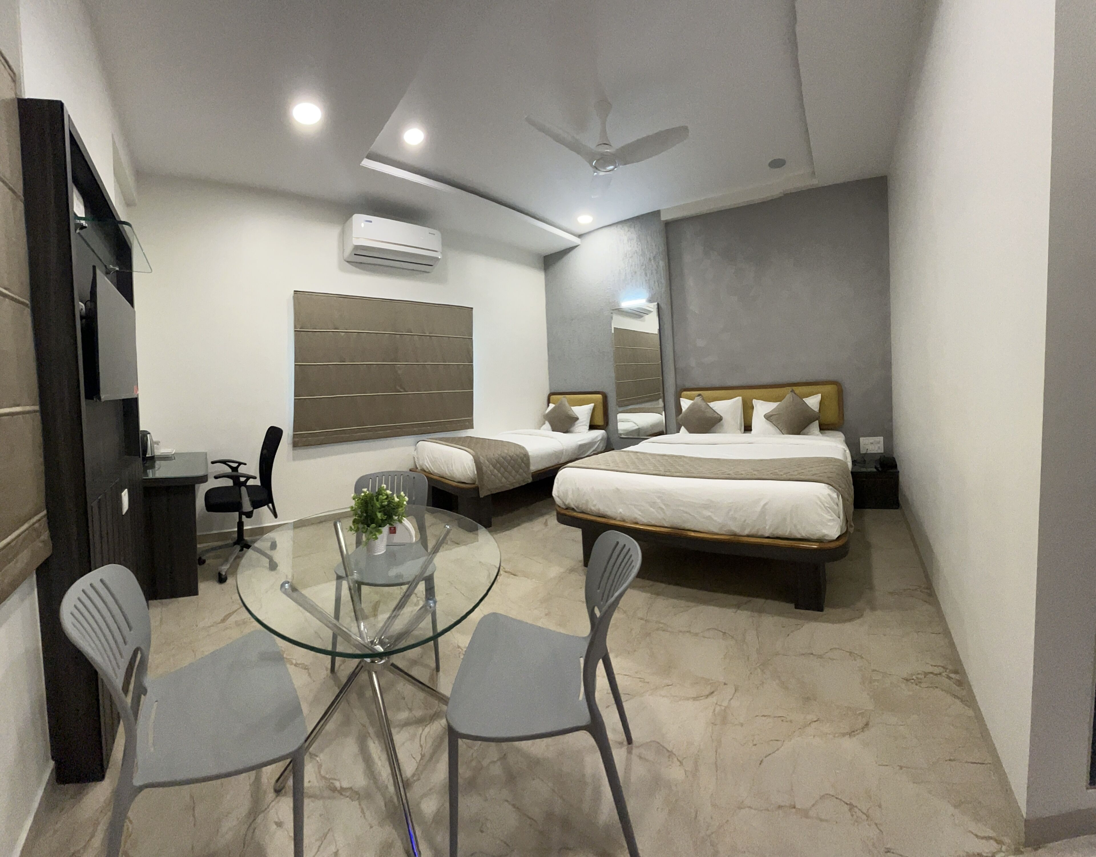 A modern hotel room with a bed, two bedside tables, a desk with a chair, a glass table with chairs, and a ceiling fan.