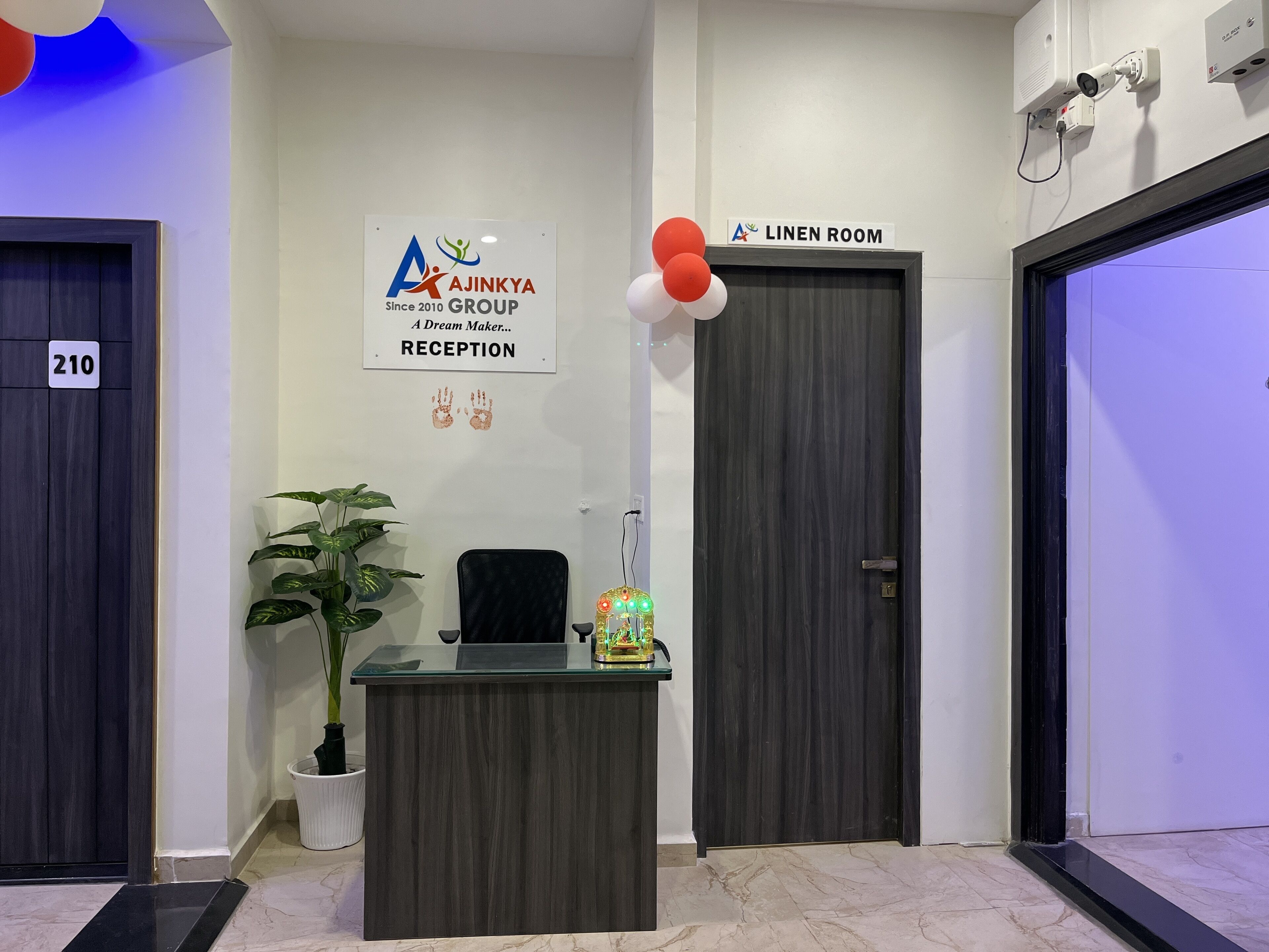 A reception area with a reception desk, a potted plant, a sign for Ajinkya Group Reception, and doors labeled '210' and 'LINEN ROOM'.