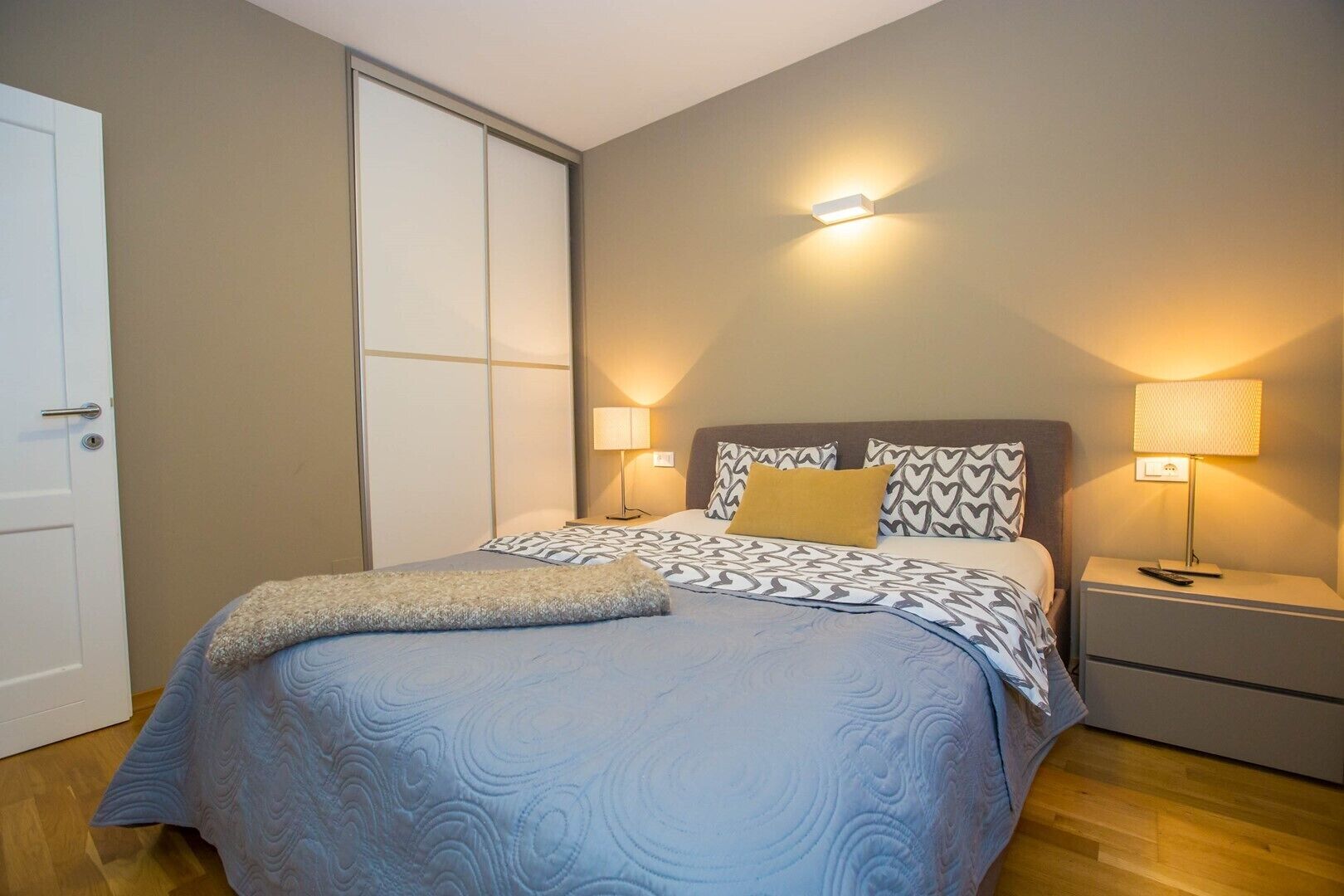 A bedroom with a bed, two bedside tables, a nightstand, a door, and a sliding closet.