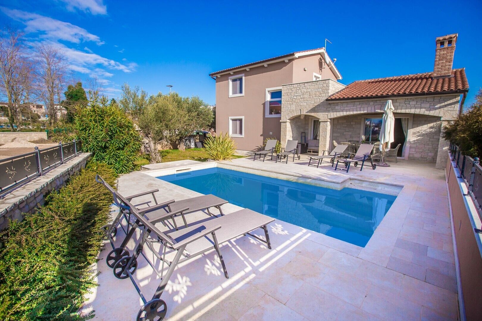 A swimming pool with lounge chairs, a stone building with a tiled roof, and a clear blue sky.