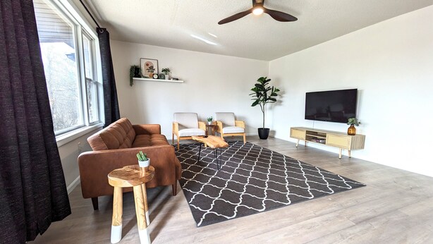 A modern living room with a brown sofa, two armchairs, a coffee table, a black patterned rug, a ceiling fan, a TV, and a potted plant.