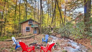 Une cabane rustique avec un petit porche, entourée d’une forêt aux couleurs automnales. On y trouve trois chaises Adirondack, de couleur rouge, bleue et blanche.