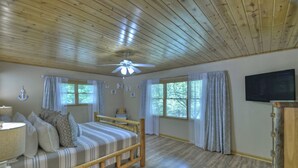 Ein Zimmer mit Holzbalkendecke, einem Bett mit Kopfteil, einem Nachttisch, einer Lampe, einem an der Wand befestigten Fernseher und Fenstern mit Vorhängen.