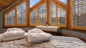 Une chambre avec un plafond en pente, des poutres en bois et de grandes fenêtres équipées de stores. Il y a deux serviettes blanches sur le lit.