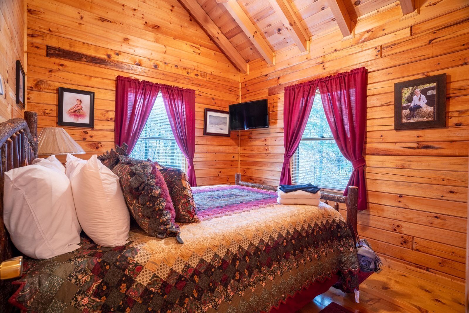 A cozy log cabin bedroom with a large bed, two windows with red curtains, and a flat-screen TV mounted on the wall.
