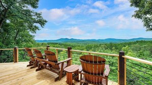 Une terrasse en bois, agrémentée de chaises Adirondack et d’une table, qui offre une vue sur un paysage verdoyant et luxuriant, avec des collines qui se dessinent à l’horizon.