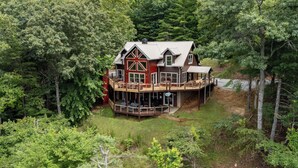 Une grande maison à deux étages, avec une imposante structure rouge qui ressemble à une grange, entourée d’une forêt dense.