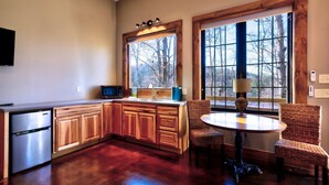 A kitchen with wooden cabinets, a stainless steel refrigerator, a microwave, and a sink. There is a round table with chairs and a lamp. Large windows provide views of trees outside. - Sugar Shack - Blue Ridge GA (Blue Ridge)