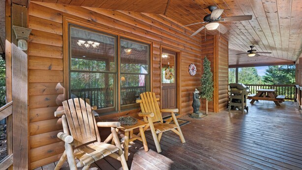 Une cabane en bois dotée d’un porche couvert, de deux fauteuils Adirondack, d’une table, d’un barbecue et d’un ventilateur de plafond.
