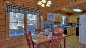 A wooden cabin interior with a dining table set for four, wooden chairs, a kitchen with wooden cabinets, and a sign that reads "on mountain time". - A Walk In The Clouds- Cherry Log GA (Cherry Log)
