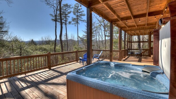 A wooden deck with a hot tub, chairs, and a table under a covered area with a view of trees and mountains. - A Walk In The Clouds- Cherry Log GA (Cherry Log)